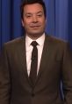 Jimmy Fallon in a dark suit, delivering a comedic monologue, backdrop featuring blue curtains. Live show atmosphere.