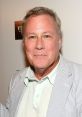 John Heard smiling at an event, dressed in light-colored attire, known for his work in film and television.