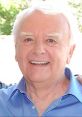 Mark Elliott, renowned Disney voiceover narrator, smiling warmly in a blue shirt, captured outdoors with a cheerful demeanor.