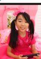 Lucy from "Lucy Toys And Colors" Season 1-2 games, smiling in a pink outfit, sitting in a colorful playroom setting.
