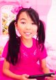 Lucy from "Toys and Colors" smiling, wearing a pink outfit with a matching bow, excitedly holding a device in her playroom.