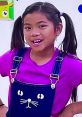Cheerful girl with pigtails wearing a purple shirt and cat-themed overalls, promoting Toys and Colors Santa's Little Genies Emma.