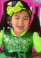 Cheerful girl wearing a sparkling green outfit and a bright bow, posing playfully in front of a colorful playhouse.