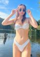 Sakura Shymko poses confidently by a serene lake, showcasing her stylish bikini and sunglasses under a clear blue sky.