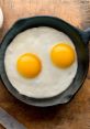 Two sunny-side-up eggs in a cast-iron skillet on a wooden surface, showcasing their vibrant yellow yolks.
