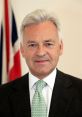 Portrait of Alan Duncan, British politician, featuring a smart suit and a light green tie, with flags in the background.