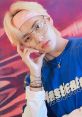 Stylish young man with blonde hair, glasses, and a headband, wearing a blue graphic tee, posing thoughtfully against a vibrant background.