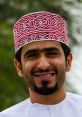 Omani man smiling, wearing traditional white dishdasha and red embroidered kumma against a blurred green background.