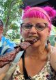 Woman with pink hair and glasses enjoying a large skewer of grilled meat at an outdoor event, showcasing a fun summer vibe.