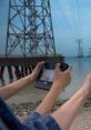 Person holding a handheld device near water, power lines visible in the background, showcasing technology in a scenic setting.