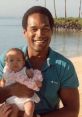O. J. Simpson smiling while holding a baby adorned with a floral lei on a sunny beach. Family moments captured.