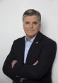 Sean Hannity poses confidently with arms crossed, wearing a blazer and light blue shirt, showcasing a professional demeanor.