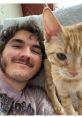 Jschlatt smiles for a selfie with a playful orange cat, showcasing a cozy, friendly moment at home.