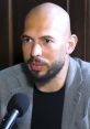 Andrew Tate engaged in a discussion, wearing a gray blazer, focusing intently on the microphone in front of him.