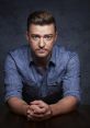 Justin Timberlake poses thoughtfully with hands clasped, wearing a blue shirt against a dark background, showcasing his charisma.
