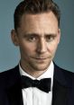 Tom Hiddleston, dressed in a classic tuxedo with a bow tie, gazes confidently against a blue background.