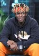 KSI in a black hoodie and orange pants, smiling while holding a microphone at an event, showcasing his vibrant style.