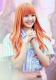 Lisa from BLACKPINK smiling happily with rosy orange hair, wearing a striped shirt, radiating K-pop charm.