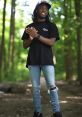 Kai Cenat smiles and poses outdoors in a forest, wearing a black t-shirt and distressed jeans, embracing nature's vibe.