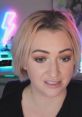 Woman with short blonde hair and makeup, engaged in a livestream with colorful neon lights in the background.