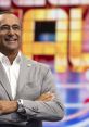Carlo Conti smiles confidently in front of a colorful game show backdrop, showcasing his charismatic television presence.