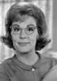 Joyce Gordon smiling with stylish glasses, showcasing a classic 1960s look and charm in a warm indoor setting.