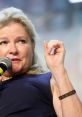 Kate Mulgrew speaking at an event, holding a microphone and gesturing with her hand, wearing a wristband.