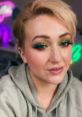 Close-up of a woman with vibrant green and gold eye makeup, wearing a grey hoodie, in a colorful, neon-lit setting.
