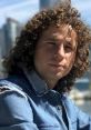 Luisito Comunica with curly hair, wearing a denim jacket, poses in a vibrant urban setting, exuding a relaxed vibe.