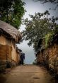 Old man walking along a rural pathway, flanked by traditional cottages and vibrant greenery, evoking tranquility and simplicity.