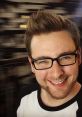 Tom Ridgewell smiling in a casual shirt, featuring stylish hair and glasses, against a blurred background.