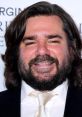 Matt Berry smiles confidently, showcasing his distinctive beard and long hair at a formal event, exuding charisma.