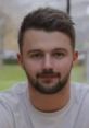 Young man with a short beard and stylish haircut, wearing a light shirt, smiling outdoors in a relaxed environment.