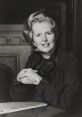 Margaret Thatcher smiling with hands crossed, wearing a black blouse, in a formal setting with elegant decor.
