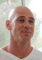 Johnny Sins smiling, showcasing his signature bald look, in a casual white shirt against a bright background.