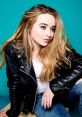 Sabrina Carpenter poses stylishly in a black leather jacket against a vibrant teal background, showcasing her signature look.