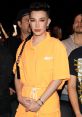 James Charles in a bright orange outfit, showcasing bold makeup and a confident pose at a fashion event.