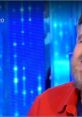 Christian De Sica smiles on stage, showcasing his comedic talent against a vibrant blue backdrop, captivating the audience.