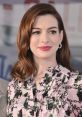 Anne Hathaway smiles warmly at a public event, showcasing her stylish floral blouse and glossy, wavy hair.