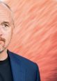 Louie C.K. in a blue striped suit against a vibrant orange background, showcasing his distinctive style and persona.