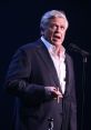 Ron White performing on stage, holding a cigar, delivering humor in a suit, showcasing his signature storytelling style.