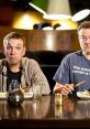 Two men at a table with food, one wearing a CinemaSins-themed shirt, sharing a moment of comedic disbelief.