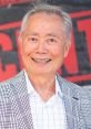 George Takei smiling broadly, wearing a checkered suit jacket, at a promotional event with a colorful background.