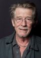 Portrait of John Hurt, renowned actor known for his impactful roles and distinctive style, smiling against a dark backdrop.
