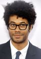 Richard Ayoade with curly hair and glasses, posing confidently in a black suit at a red carpet event.