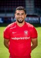 Mokhtar in a red FC Twente jersey, smiling on the pitch, showcasing his passion for football and team spirit.
