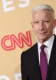 Anderson Cooper in a sharp suit, posing confidently at a CNN event with a vibrant background.