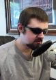 Gianni Matragrano performing a Duke Nukem impression, wearing sunglasses and speaking into a microphone.