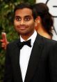 Aziz Ansari smiling in a black tuxedo with a bow tie at a glamorous event surrounded by greenery.