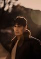 Young man in a stylish jacket with fur collar, gazing into the distance against a soft, golden sunset backdrop.